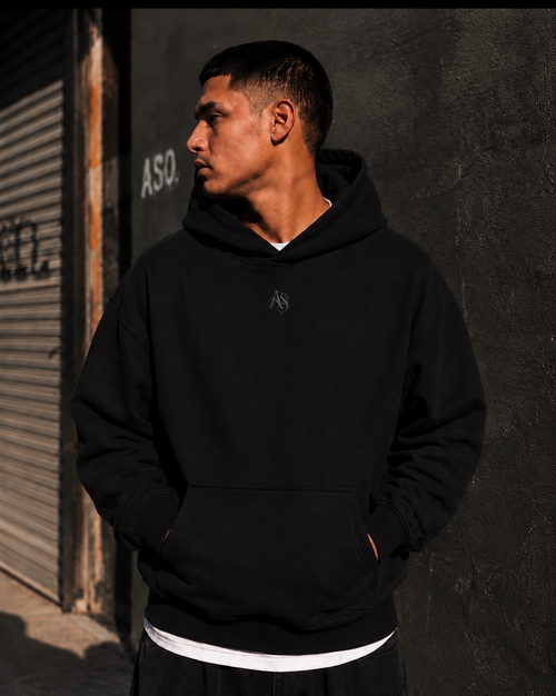 A man wearing a black wearing the black Así Soy Originals Signature Hoodie with a small embroidered logo on the chest and kangaroo pocket